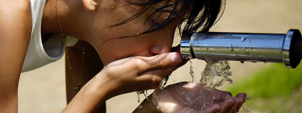 a young hispanic girl drinking from an outdoor water faucet in the heat