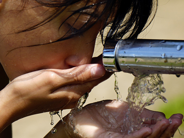 a young hispanic girl drinking from an outdoor water faucet in the heat