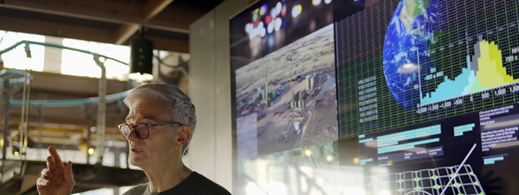 A man conducting a meeting with the aid of a large screen displaying data and photos of low carbon electricity production with solar panels and wind turbines, juxtaposed with conventional fossil fuel production